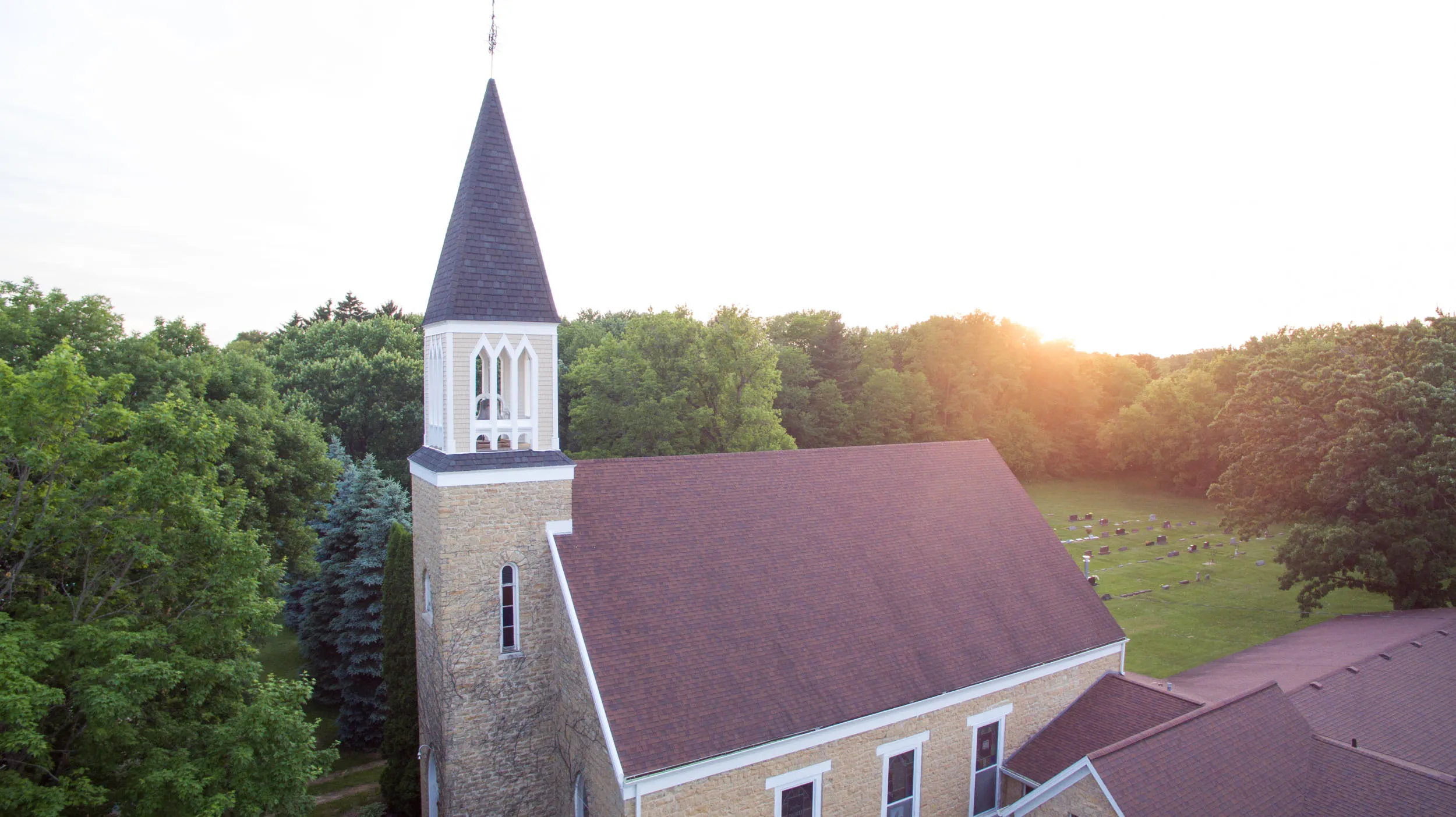 Stone church with tall steeple and brown roof surrounded by green trees and a nearby graveyard at sunset.